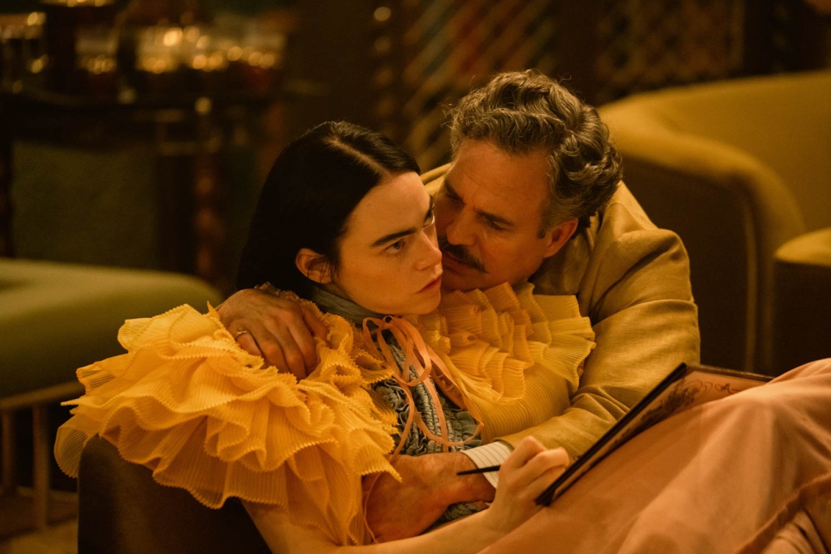 A woman in a pink, ruffled dress is sitting down and staring ahead, while a man with gray hair and mustache is hugging her from the side and leaning towards her face.