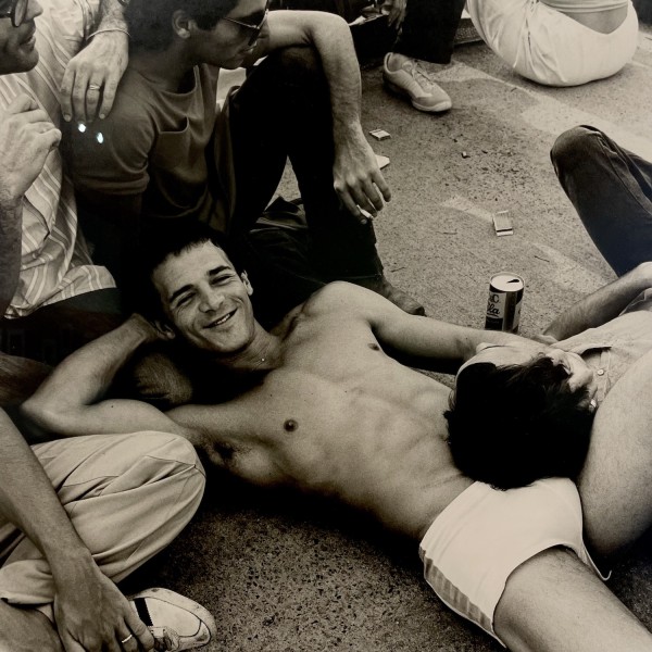 A black-and-white photo of a group of men sitting and lying down on concrete. One of them is shirtless and smiling. His head is resting in another man's lap.