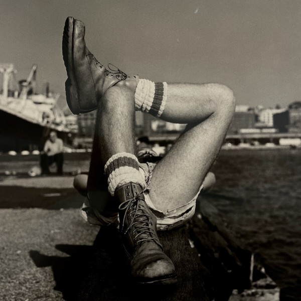 A black-and-white photo of someone lounging on a pier. He is wearing boots and one of his feet is resting on his knee.