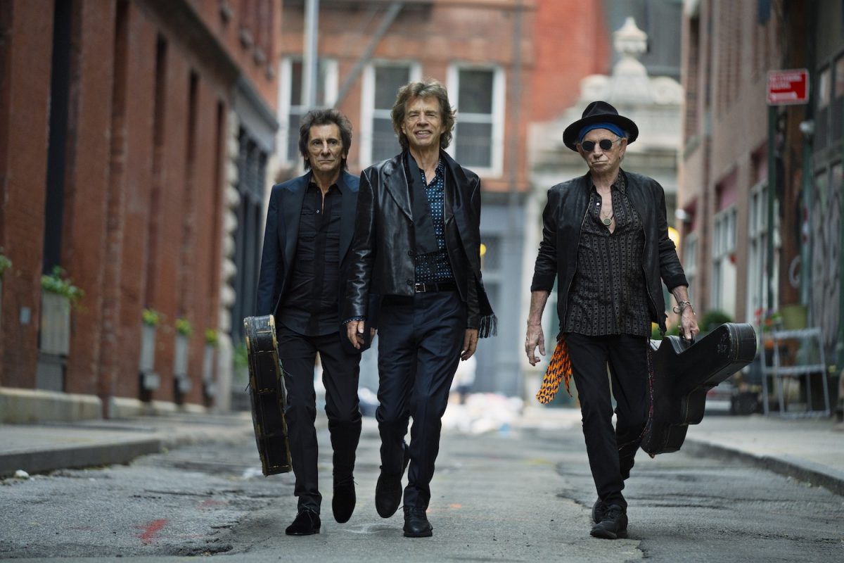 Three men walking in an alley. Ronnie Wood, the man on the left, is holding a black guitar case. Mick Jagger is in the middle. Keith Richards, the man on the right, is holding a black guitar case and has black glasses on.