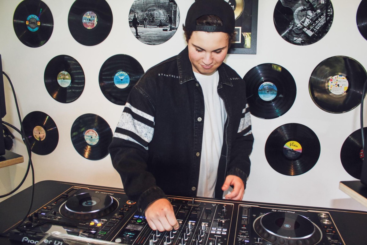 A man wearing a backwards cap, a white shirt and a black jacket stands behind a D.J.’s turntable. Behind him is a wall decorated with records.