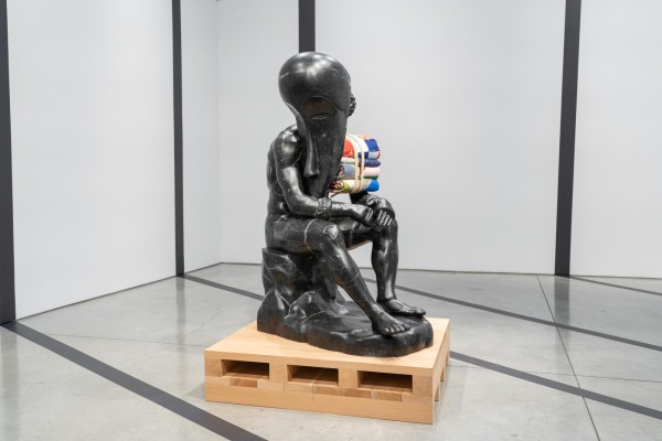 A statue made of black material of a person sitting on a rock. An African Fang mask covers one side of the person, while a bundle of quilt rests on the other side.