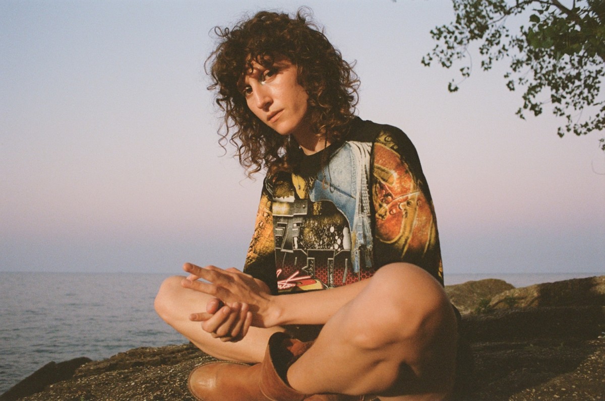A person sits on a rock above the water wearing a multicolored top and brown boots.