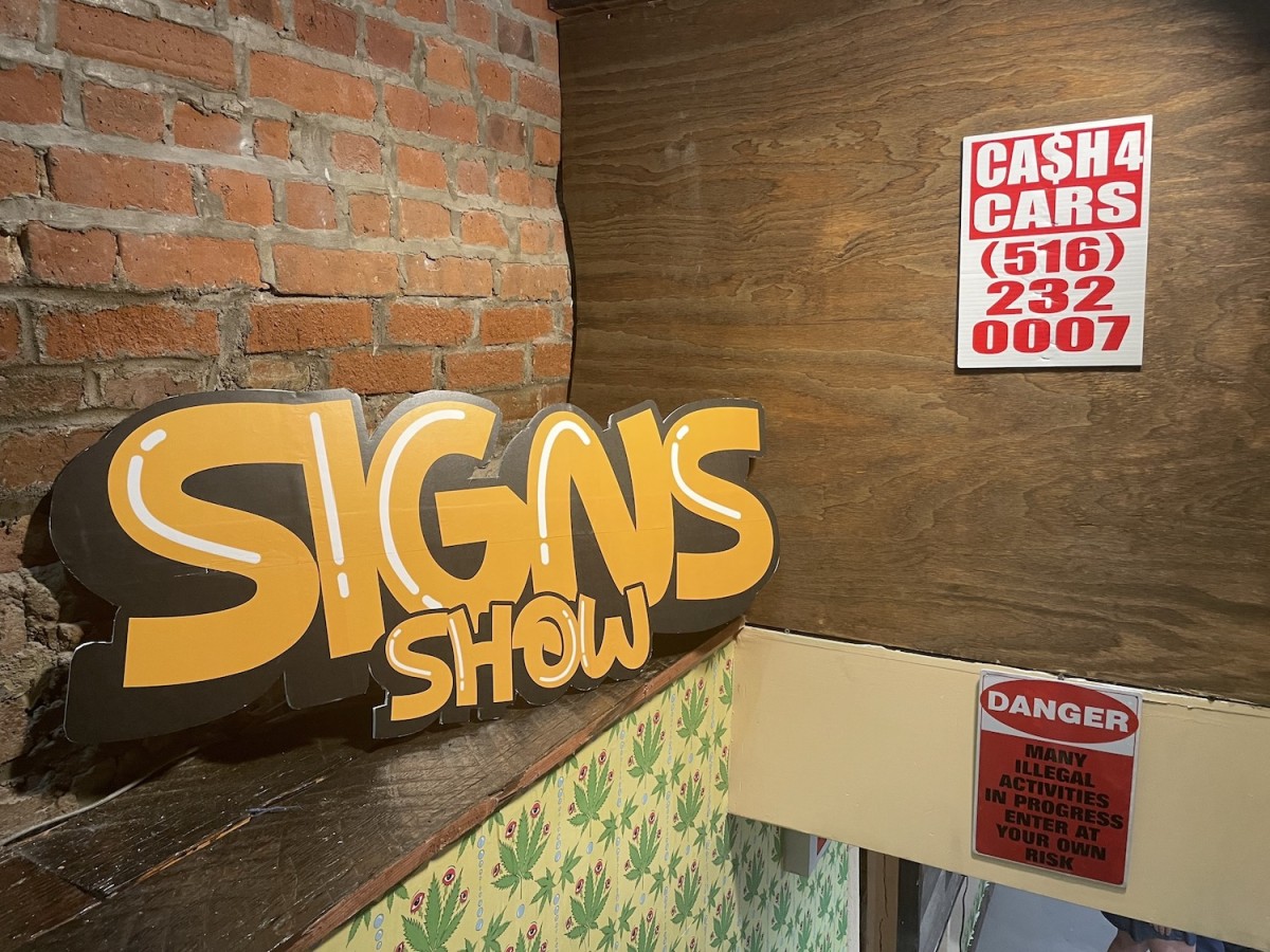 A yellow sign that reads, ‘SIGNS SHOW’ is leaning against a brick wall, above a stairwell. On the wall to the right of the sign hang two red signs.