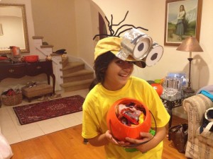 A girl in a yellow shirt and a yellow hat with big eyes holding an orange pumpkin basket full of candy, dressed as a minion from the “Despicable Me” franchise.