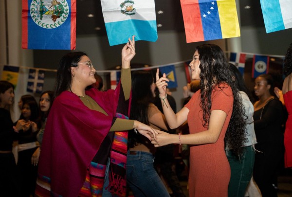 Two women dancing while holding hands. Above them hangs a row of Hispanic country flags.