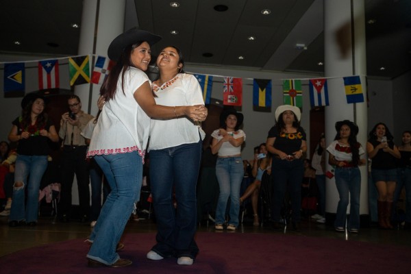 Two women dancing in a ballroom with a group of people standing behind them. Above the crowd hangs a row of Hispanic country flags.