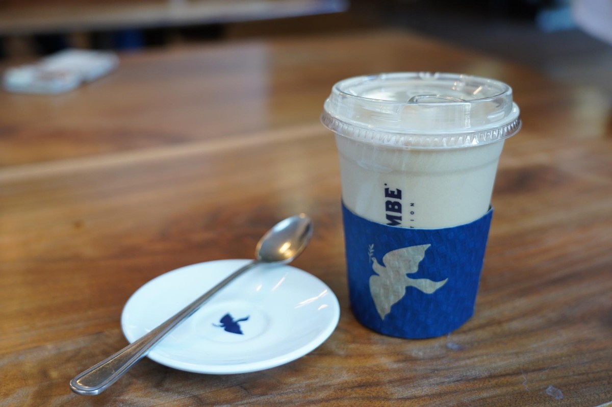 A plastic cup with a blue sleeve holding iced coffee sits on a wooden table next to a small white plate with a metal spoon resting on it.