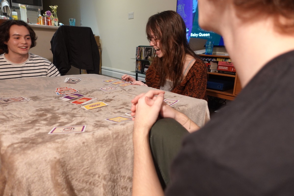 Three people are sitting around a table with a beige tablecloth and playing a card game. There are cards laid out on the table.