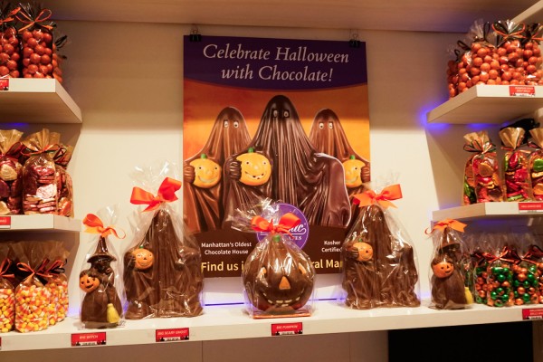 A shelf filled with various halloween themed candies including chocolates shaped as pumpkins and ghosts. At the back of the shelf is a poster reading “Celebrate Halloween with Chocolate!”.