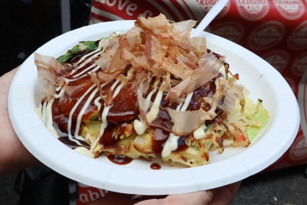An okonomiyaki, a Japanese pancake, placed on white plate with a white utensil. Red fabric with the Oconomi logo in the background.