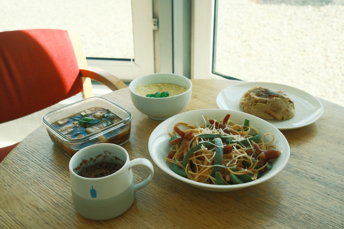 Two plates, a bowl, a glass container, and a mug on a wooden table. One plate has spaghetti, one has French toast, the bowl has Korean steamed eggs, the glass container has coke-braised pork, and the mug has a chocolate cupcake inside.