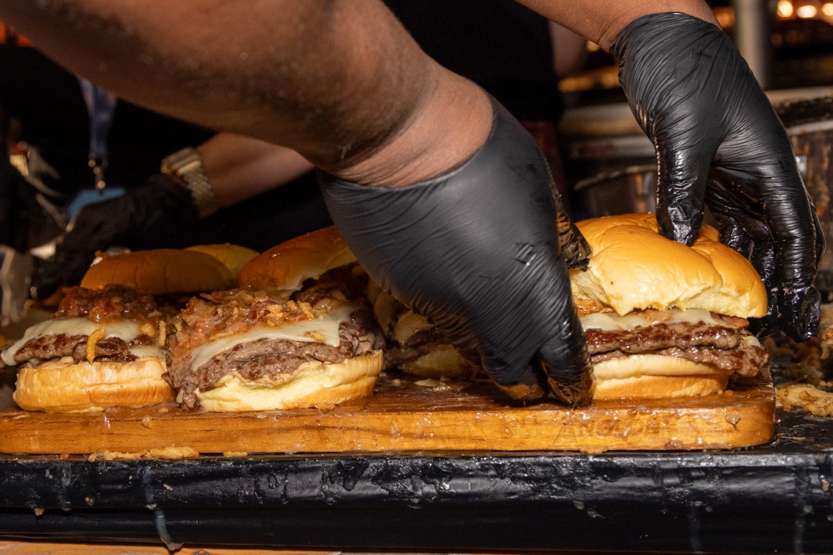A person with black gloves on picks up a burger from a brown cutting board.