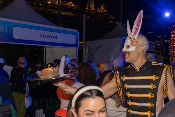 A person in a white bunny mask and black and gold sleeveless top holds a burger from the Nowon booth.
