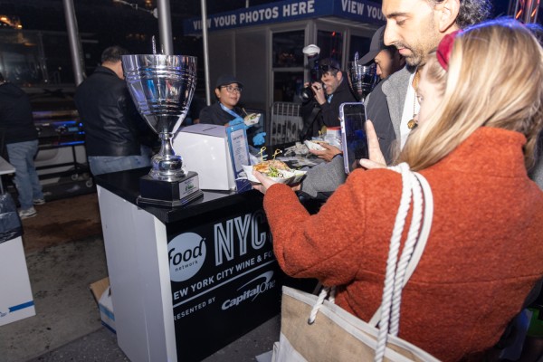 A person in a red jacket with blonde hair takes a photo of their burger with last year's silver colored trophy won by San Matteo.