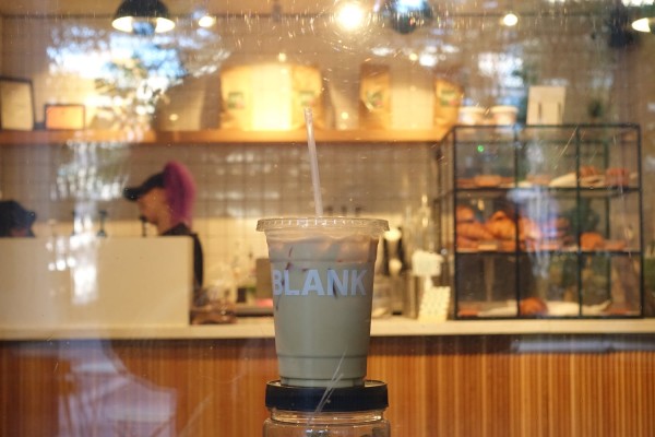 A plastic cup of ice coffee, with ‘BLANK,’ written on it in white letters. There is a glass window behind the cup and through the glass is the inside of a coffee shop.