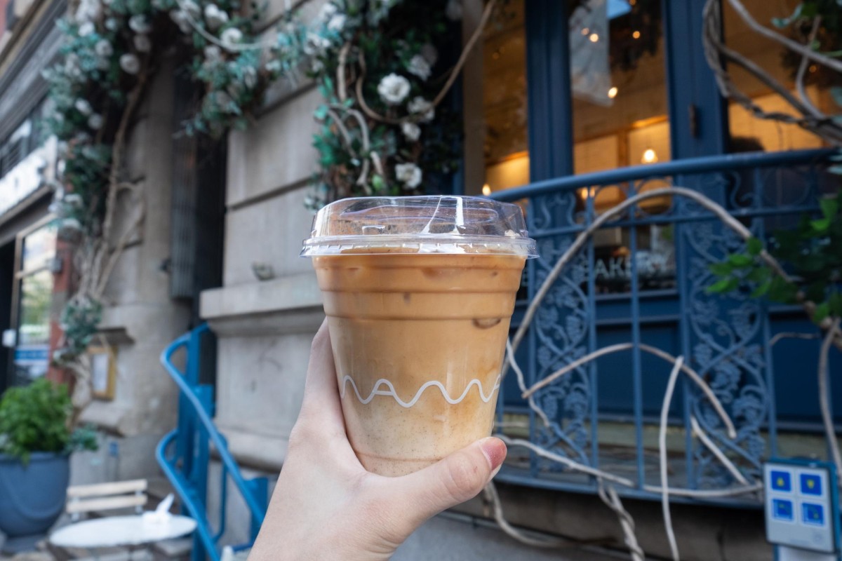 A hand holding a plastic cup of ice coffee with a white wavy design across the top of the cup that reads maman. In the background is a blue and white storefront.