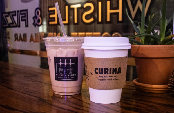 One ice coffee and one hot coffee next to each other on a brown table, with a plant to the right of them.