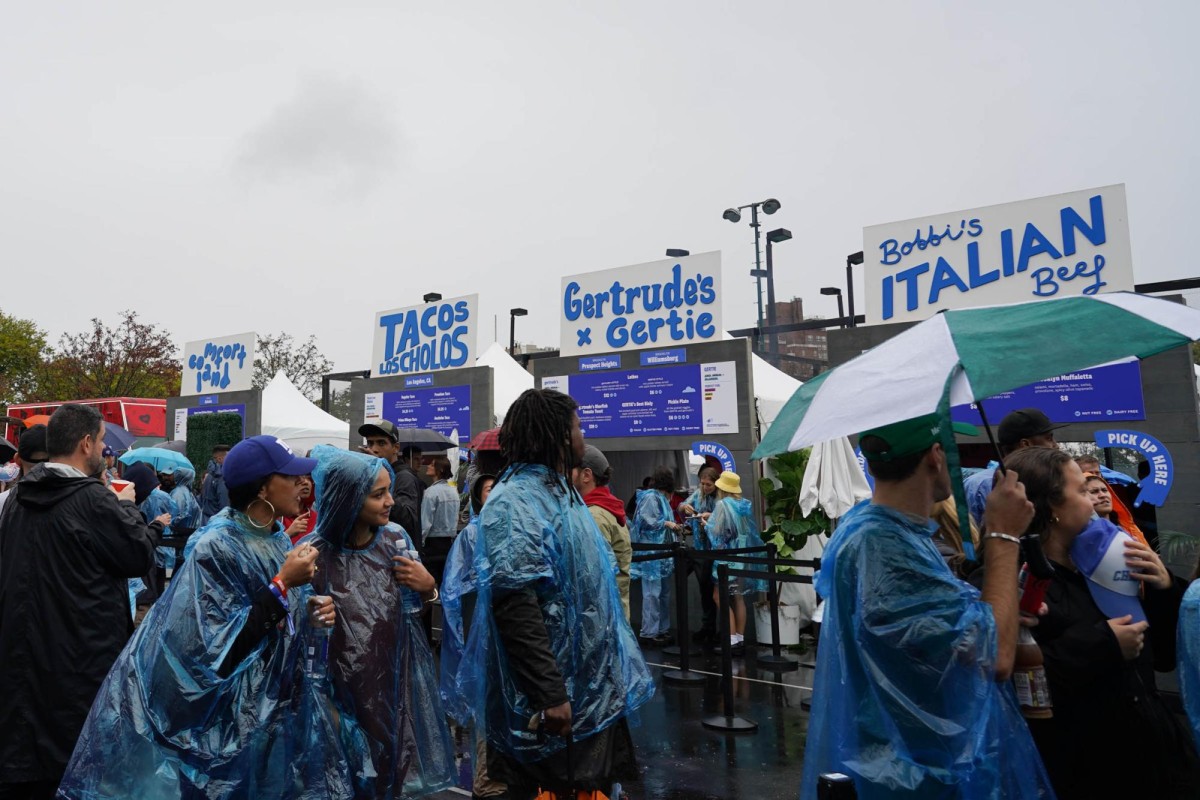 People holding umbrellas and wearing blue, plastic rain ponchos are lining up in front of food stands with blue signs that have the restaurant names written on them.