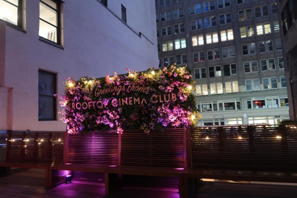 A board that says “Greetings from ROOFTOP CINEMA CLUB” decorated with flowers stands behind a row of benches on a rooftop.