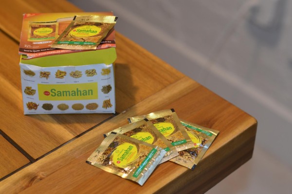 A box of tea reads “Samahan” and features pictures of the variety of spices in the tea. The tea bag packets that are spread on the same surface are gold in colour and feature the text “Samahan” and “100% Natural”.