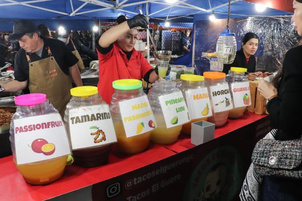 A woman standing behind jars of beverages on Tacos El Guero’s stall, while pouring a drink into a plastic cup in a plastic cup with a large spoon.