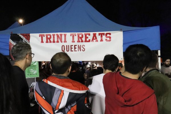 People standing in front of a tent with a white sign that says “TRINI TREATS QUEENS”