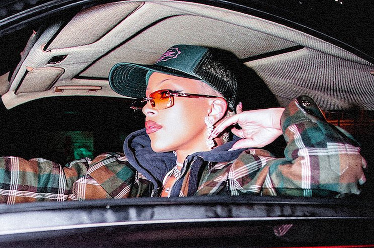 A woman in a camo-colored plaid jacket wearing a green baseball cap and sunglasses sits behind the wheel of a car.