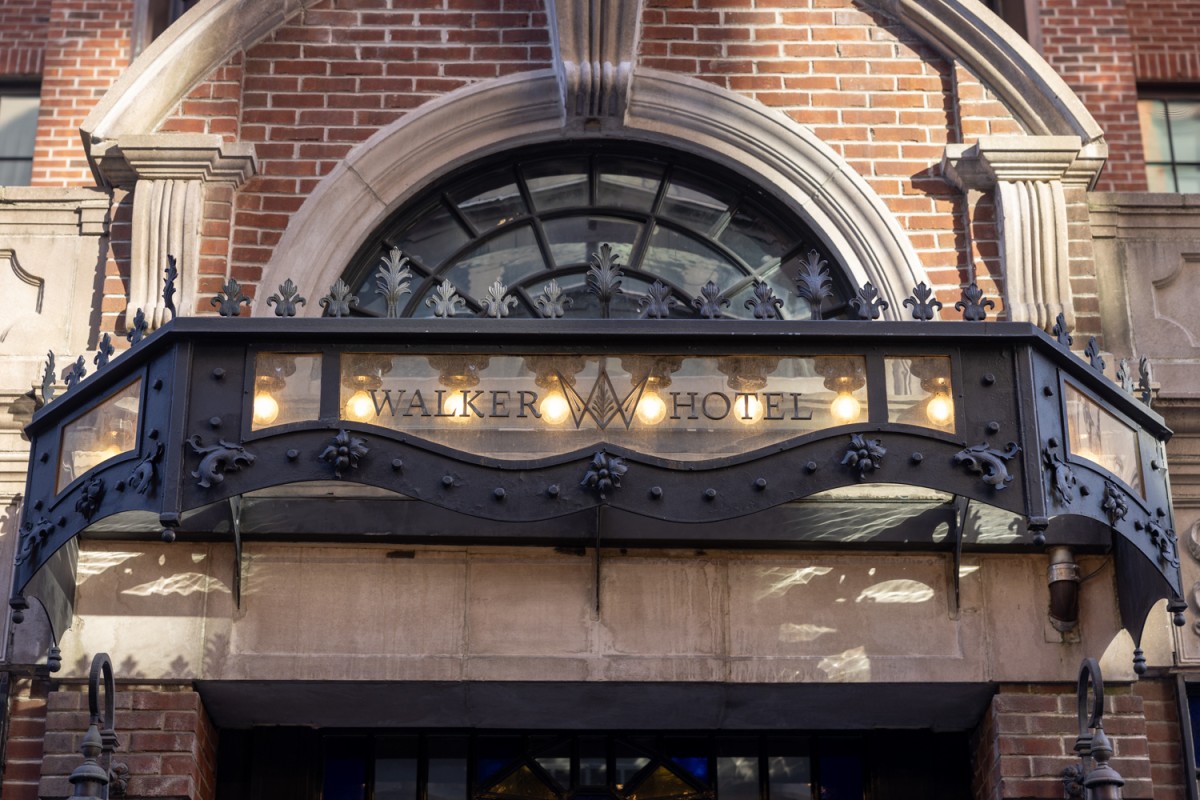 A glass sign that says “Walker Hotel” illuminated by yellow light bulbs above the entrance of the Walker Hotel.