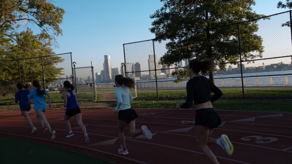 Five people are running on a track. There is grass next to them, as well as the river and the skyline.