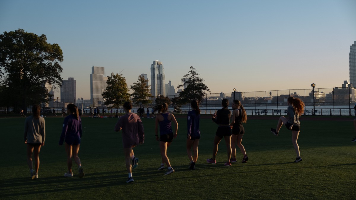 Eight people stand in a line on a grass field and are doing warmup exercises, with a few of them talking to each other.