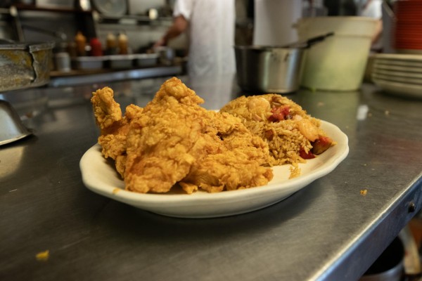 A white plate with fried chicken and fried rice on it. There are kitchen appliances and a chef in the background