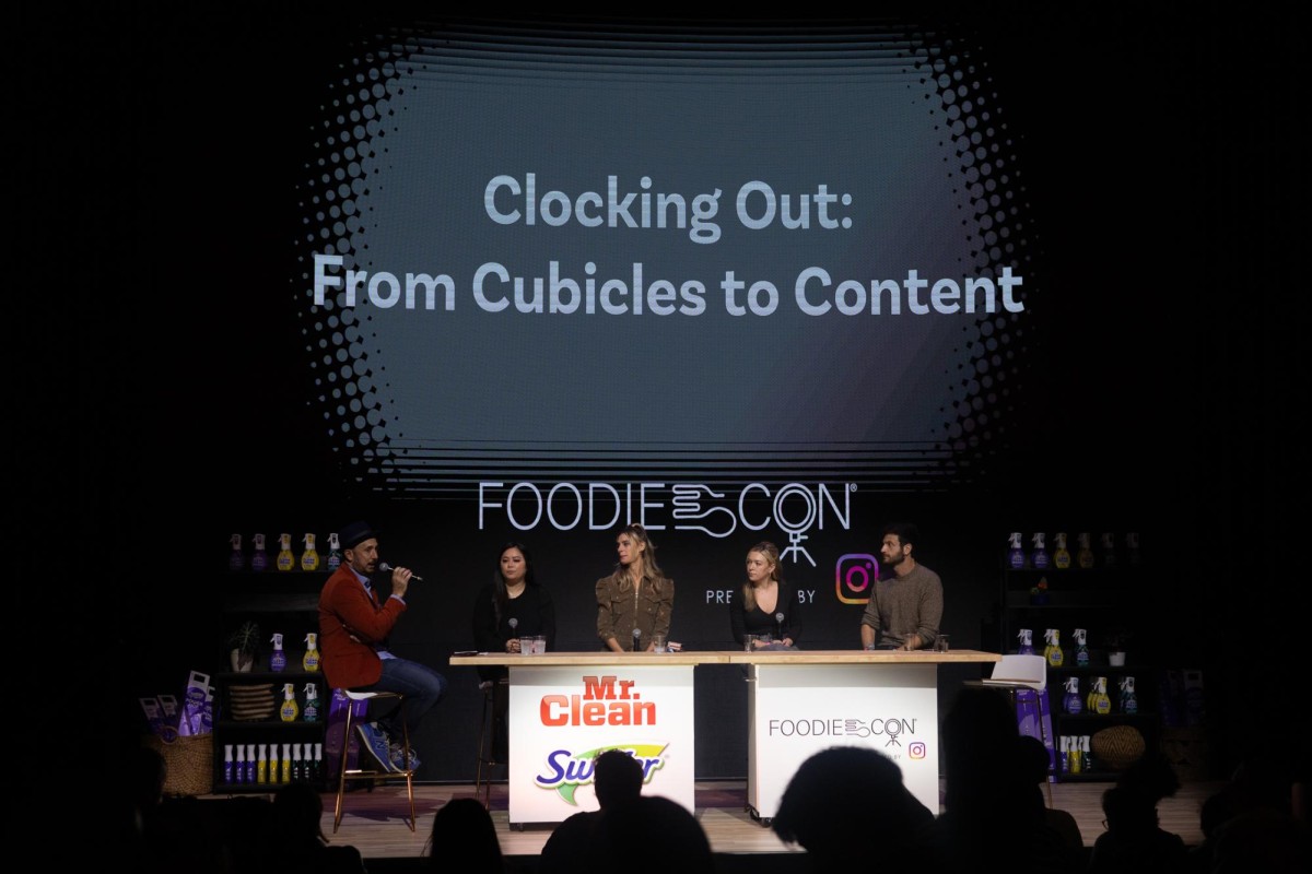 An interviewer holding a microphone is asking four panelists a question on a stage. The panel is named “Clocking Out: From Cubicles to Content.”