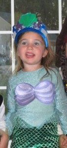 A girl in a sea green shirt with purple shells and a green tail wearing a blue headpiece with a green shell, dressed as Ariel from the Disney movie “The Little Mermaid.”