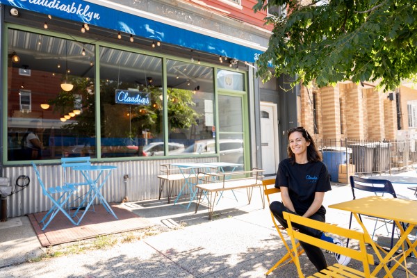 To the right of the photo is Guinevere, a co-owner of Claudia’s by Clo Cafe, sitting on a yellow chair, wearing a black t-shirt with ‘Claudia’s’ written on the top right corner. Behind her there is a blue store sign and a blue neon sign that reads ‘Claudia’s.’