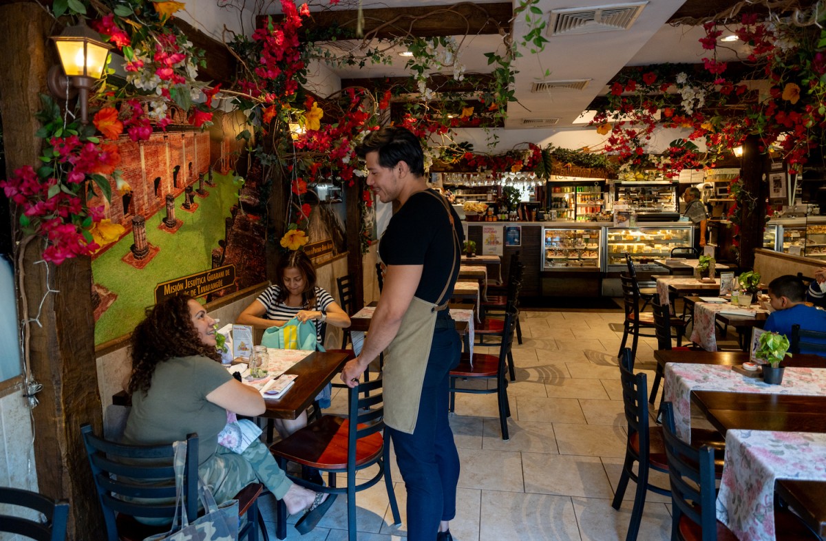 The interior of I Love Paraguay Restaurant. Two people are sitting across from each other. Jaime Issac S., a server at I Love Paraguay, is talking to them. The wall behind them is painted and there are pink flowers across it, as well as across the ceiling.