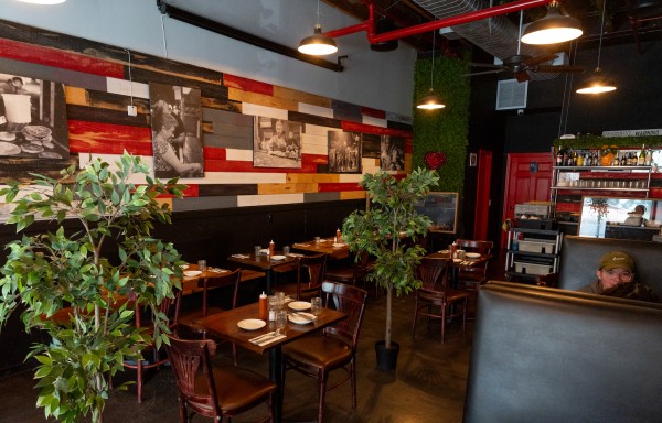 The interior of Arepa Lady. There are dark brown, wooden chairs and tables. There are black and white photographs on the wall, and two plants in pots. To the right of the photo there is a man wearing a brown cap sitting at a booth.
