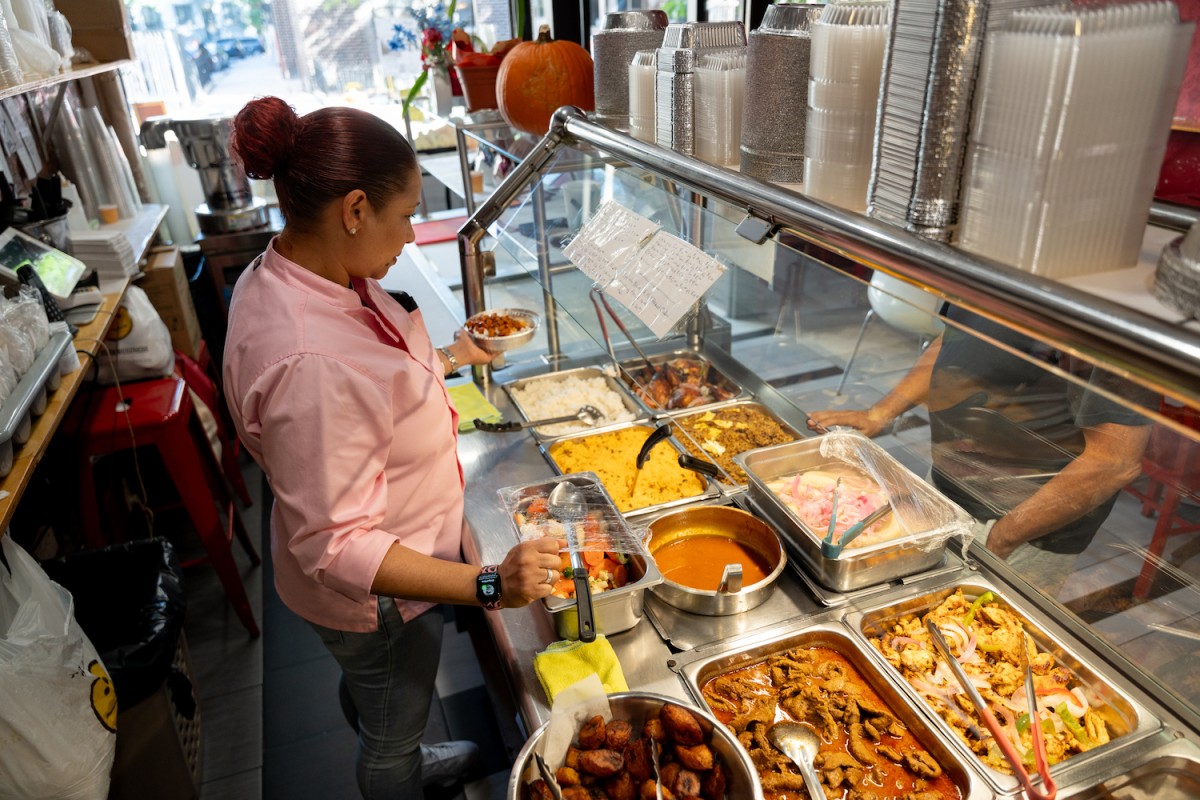 Rossy Caba is preparing an order at Rossys Bakery & Coffee Shop. She is wearing a pink shirt and holding up a container with food. She is standing behind a buffet full of different dishes.