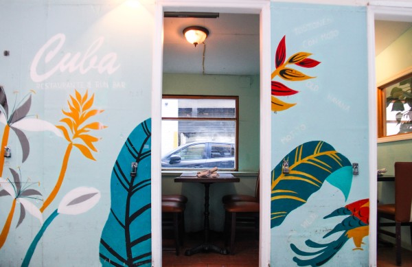 The outside seating area for the restaurant Cuba, with blue and orange leaves drawn on the walls and the name “Cuba” printed on the walls, as well as chairs and a table outside.