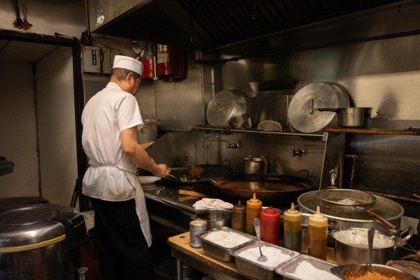 A man in a white t-shirt, a white hat and a white apron is cooking with his back turned. There are silver cooking appliances around him and a variety of sauces to his right.