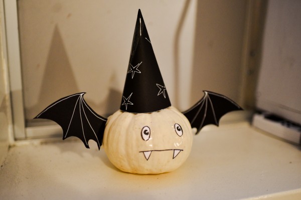 A white pumpkin decorated with a black pointy hat with stars, a pair of black wings, a pair of eyes, and a mouth with two fangs.