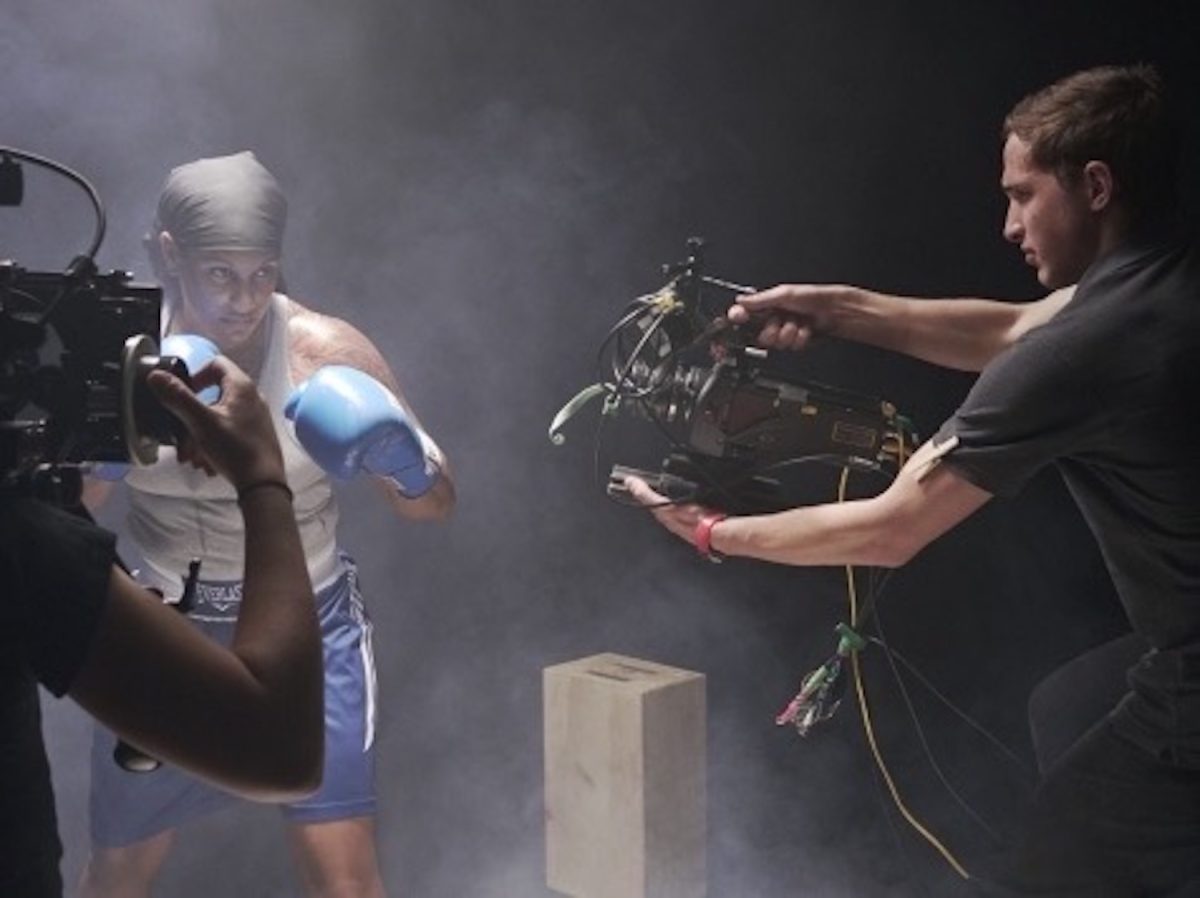 A man in gray holding a camera facing a person with boxing gloves.