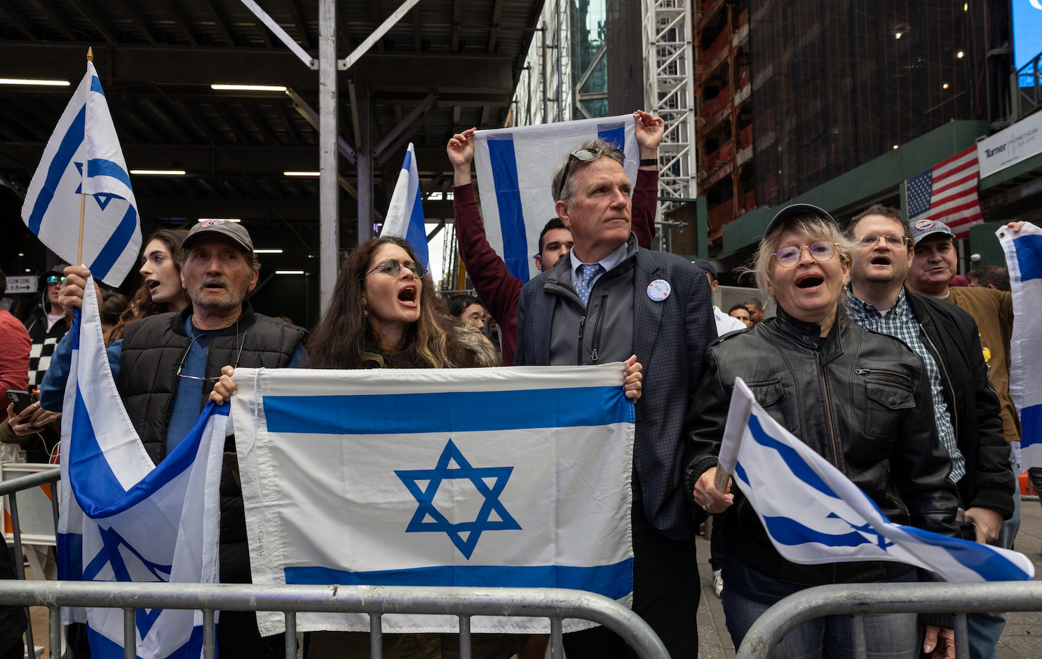 A group of people standing behind a fence shouting while holding various-sized Israeli flags.