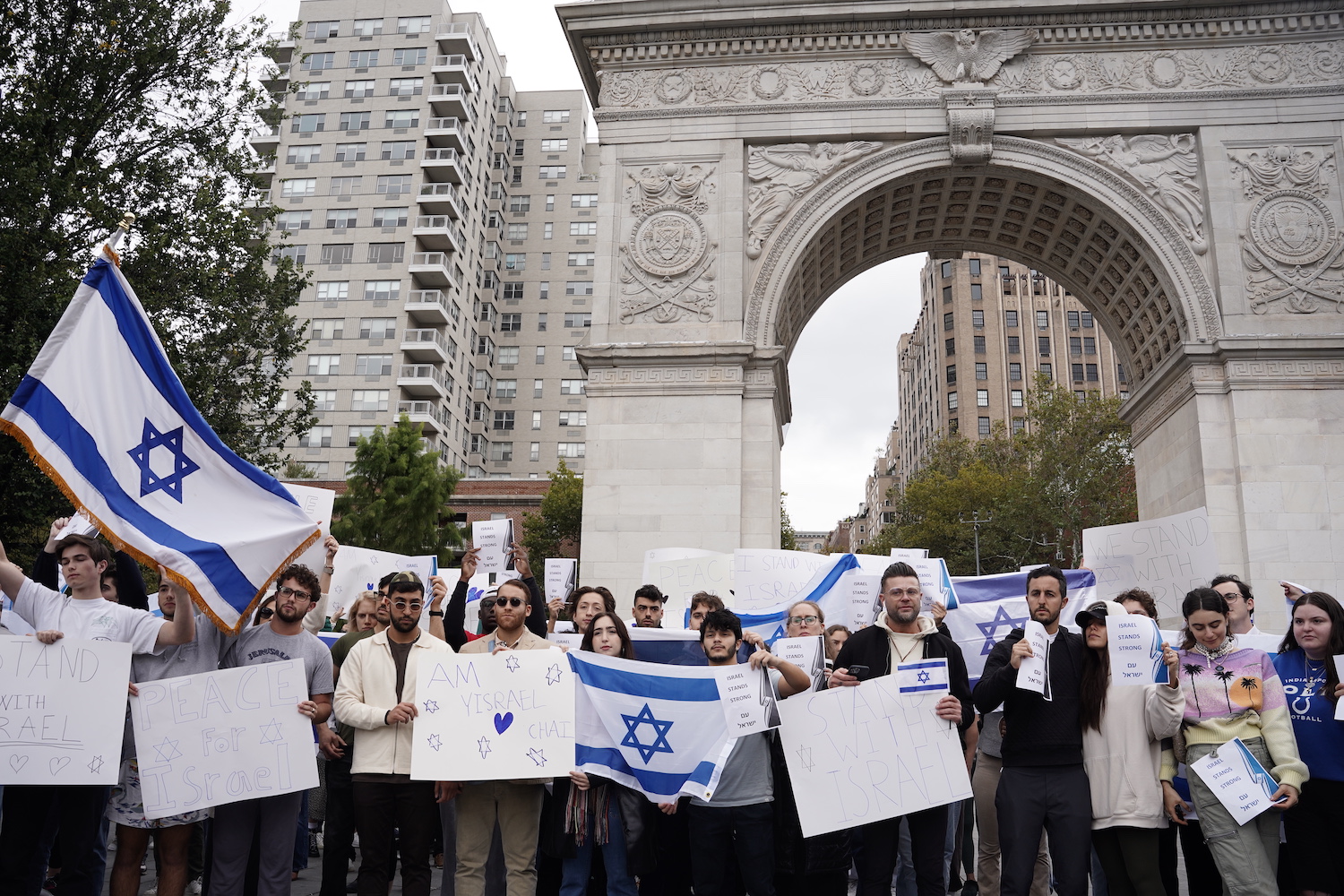 A crowd of people with signs and flags stand in front of the Washington Square Arch.