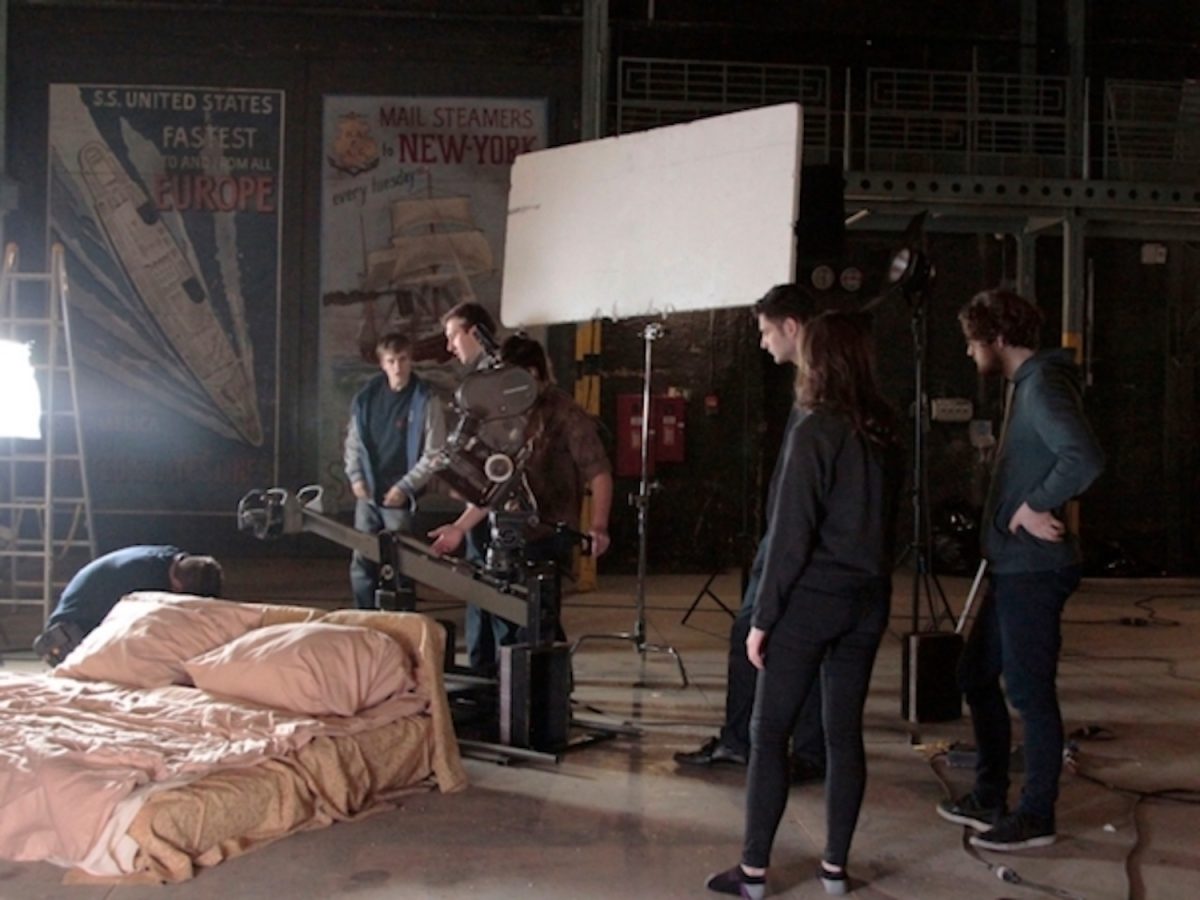 A+group+of+students+standing+in+front+of+a+beige+mattress+with+various+pieces+of+film+equipment+in+a+warehouse.