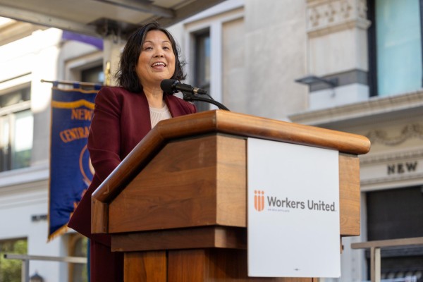 A woman wearing a dark red jacket speaks behind a "Workers United" podium.