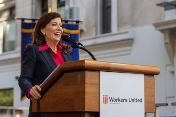 A woman wearing a black jacket and a red shirt speaks behind a podium with the sign “Workers United” on it.