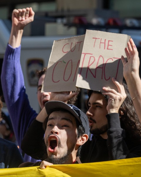 A man wearing a cap yelling behind a yellow banner, behind him is a man holding a sign that says “Cut the Contract”.