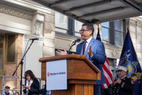 A man wearing a light blue blazer and a white shirt speaks with his arms open behind a podium with a “Workers United” sign on it.