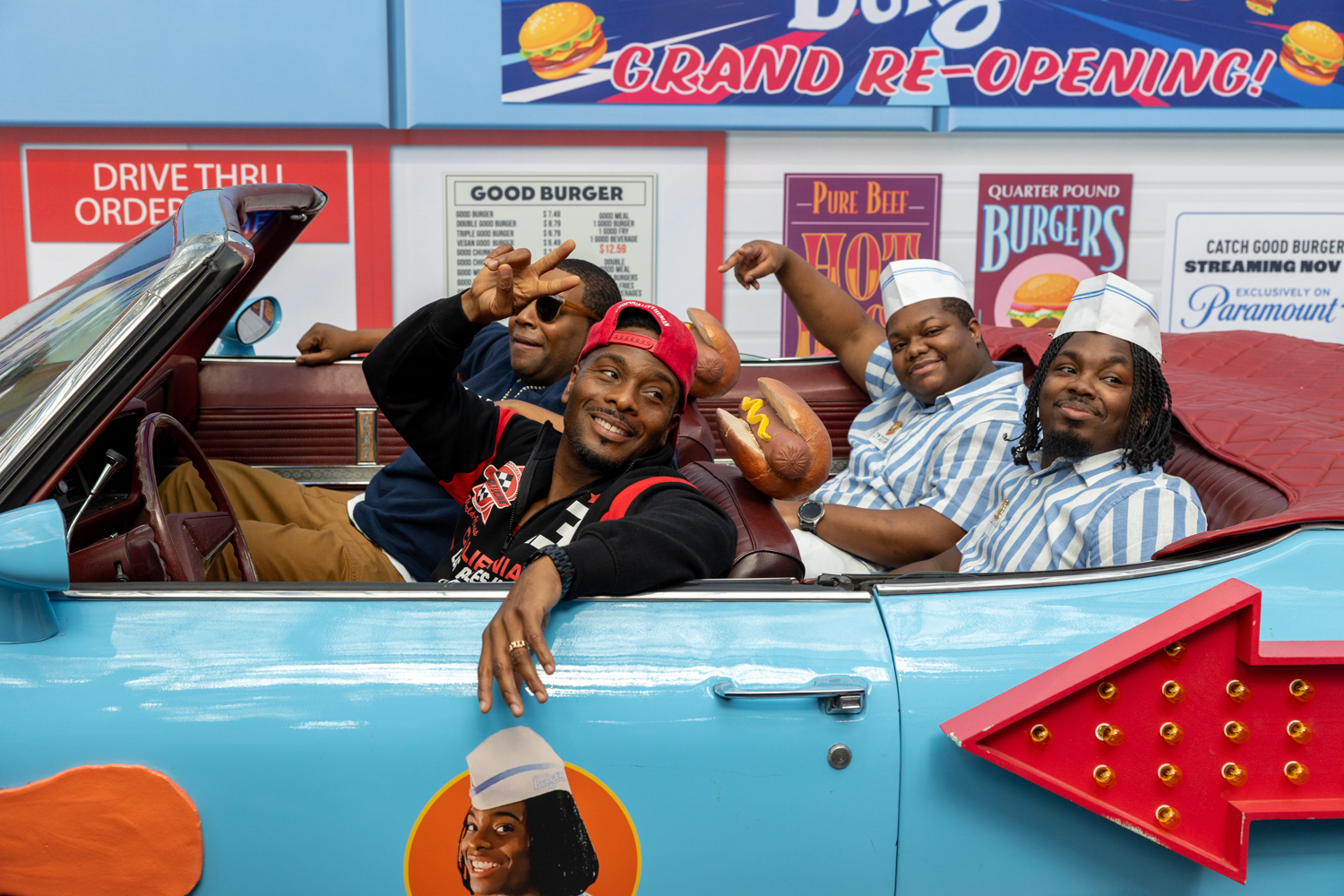 Four people sit in a blueand red convertible dressed as characters from the television show “Good Burger”. Behind them is a wall painted to look like a fast food drive-thru window complete with “Good Burger” posters.
